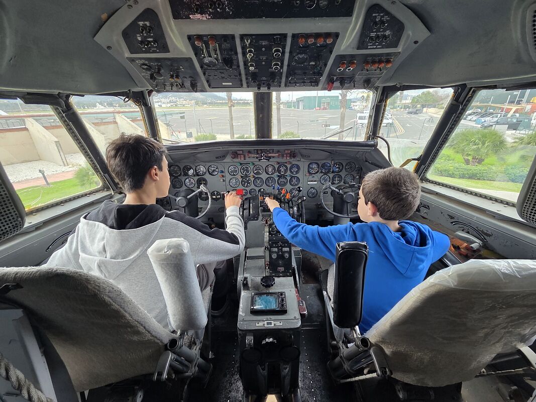 Nico y Bruno sentados en los dos asientos de piloto de la cabina de un avión real, con los cuadros de relojes delante y la pista al otro lado del cristal