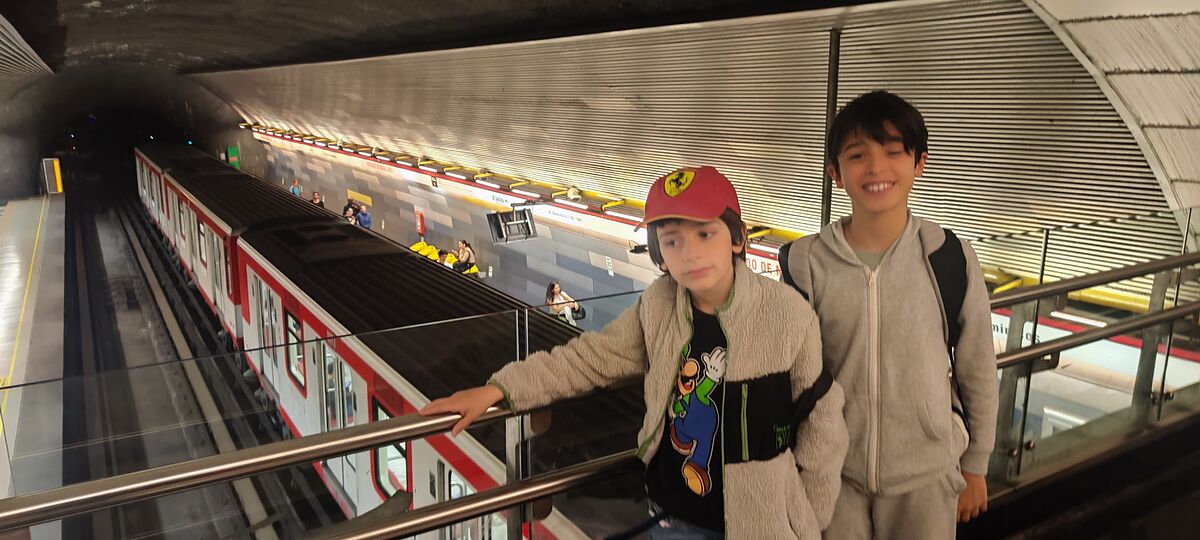 Nico y Bruno sobre el andén del metro de Santiago con un tren rojo y blanco entrando en la estación