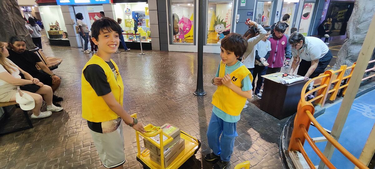 Nico y Bruno con chalecos amarillos empujando un carro lleno de paquetes por la calle de KidZania