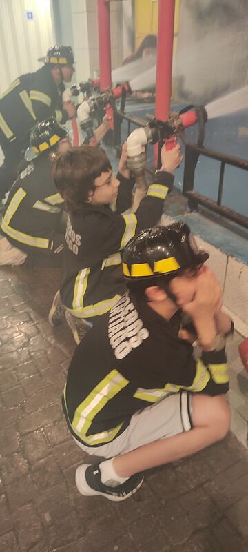 Nico y Bruno vestidos de bomberos con casco, manejando una manguera en el simulacro de incendio