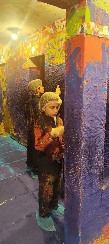 Nico con mono, gorro y calzas pintando una pared cubierta de manchas de colores