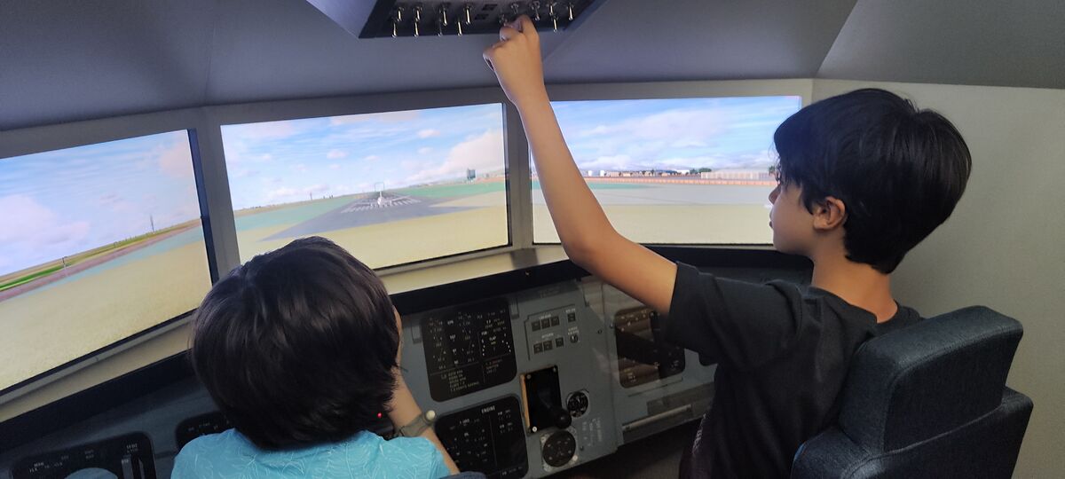 Nico en el asiento de piloto de un simulador de vuelo accionando los mandos del techo, con la pista de aterrizaje en las pantallas