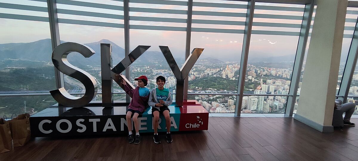 Nico y Bruno sentados en las letras del mirador Sky Costanera con Santiago y la cordillera al atardecer detrás del cristal