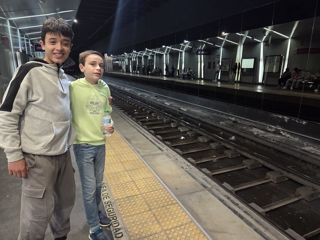 Nico y Bruno en el andén de una estación de cercanías, junto a la línea de seguridad amarilla